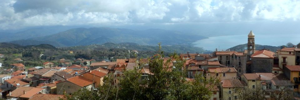 Ein Dorf hinter mehreren Bäumen und einem großen See im Hintergrund - gesehen auf einer individuellen Wanderung in Süd-Italien